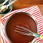 Paleo homemade bbq sauce in a small pan on a red and white striped towel with a small whisk in the pan.