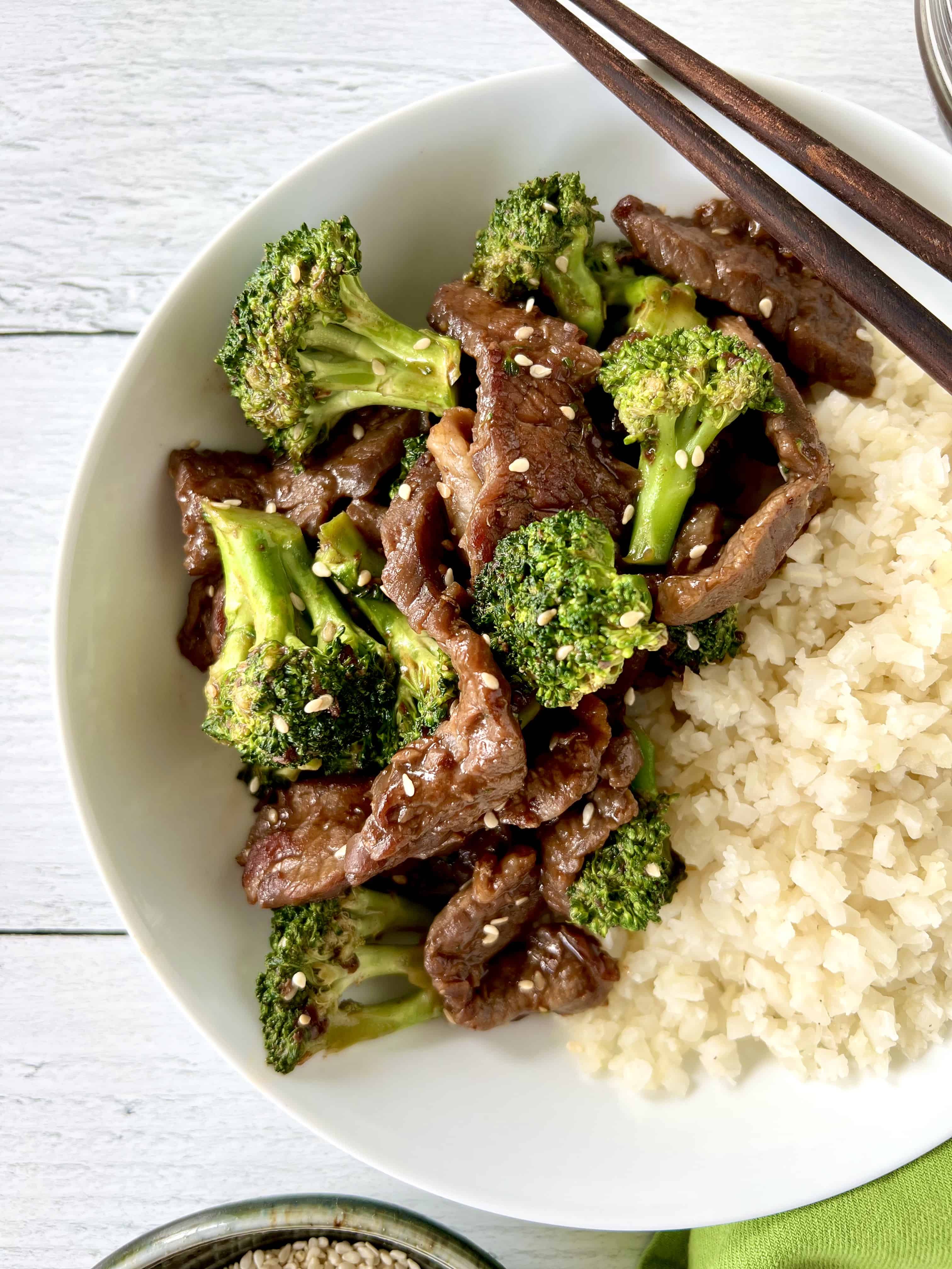 Whole30 beef stir fry in a white bowl with cauliflower rice and chopsticks.