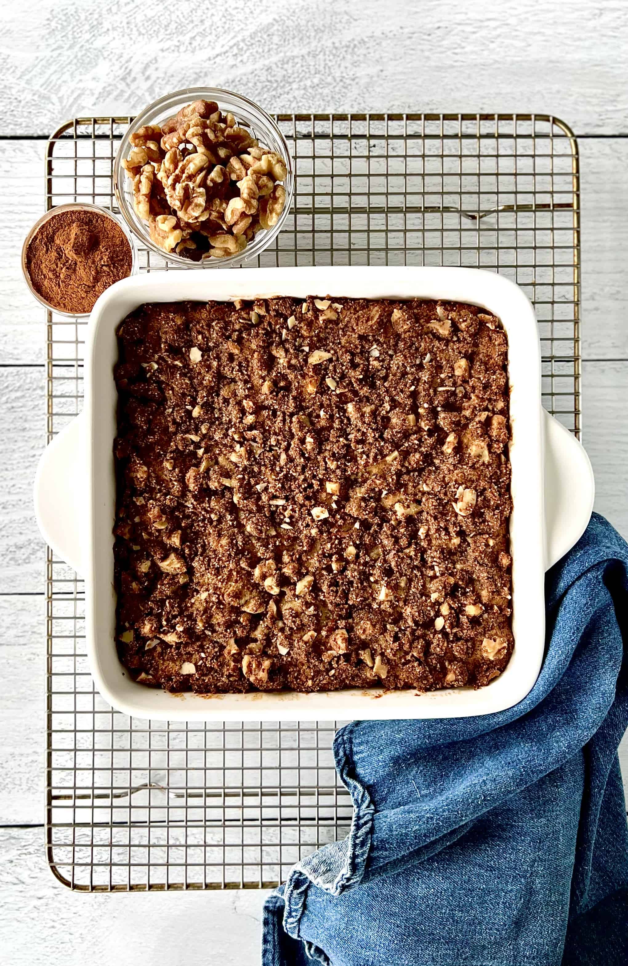 Healthy coffee cake on a cooling rack with a blue towel and small bowls of cinnamon and walnuts.