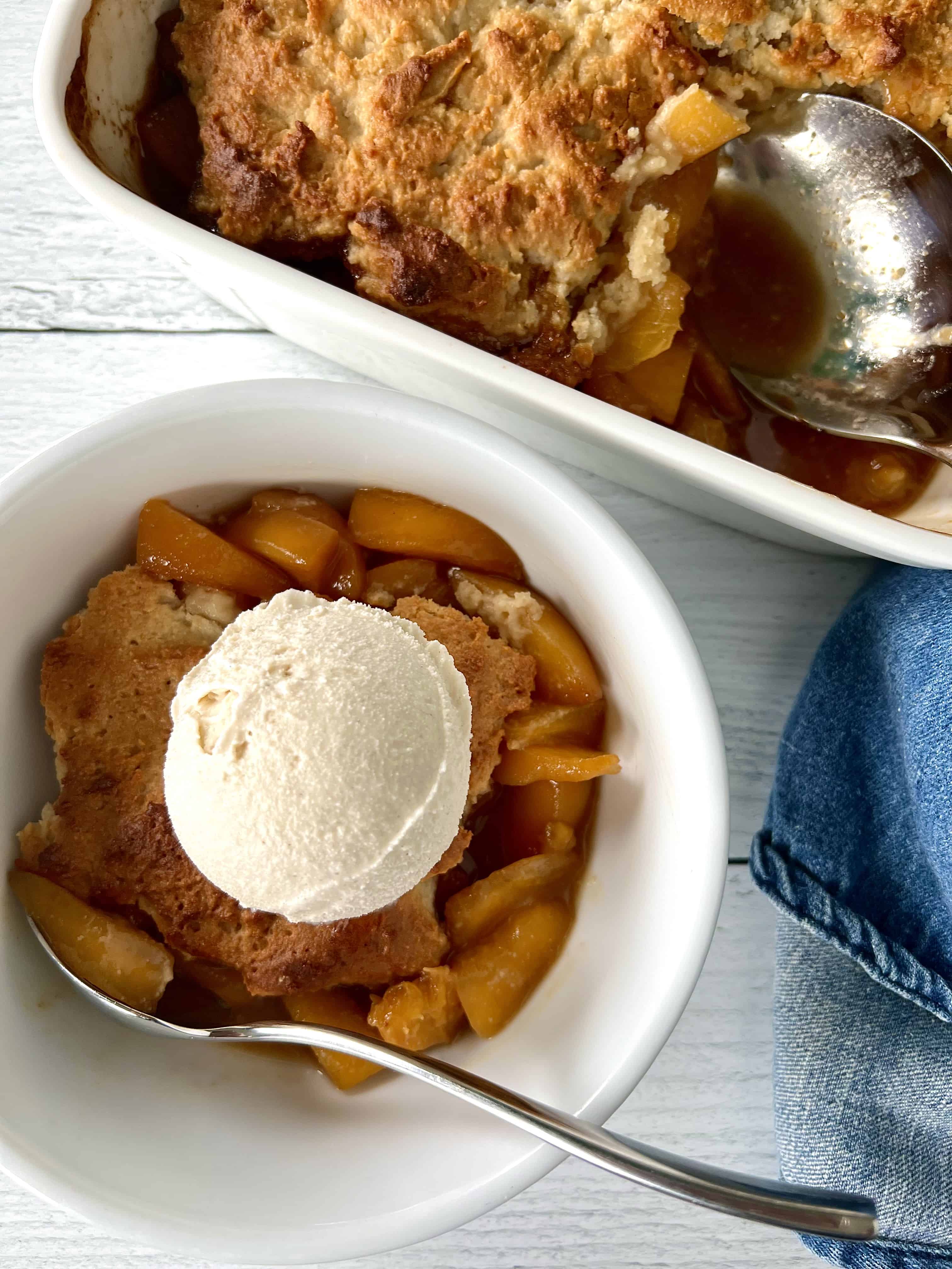 Healthy peach cobbler in a white square baking dish with a metal serving spoon and in a white bowl with ice cream on top and a spoon.