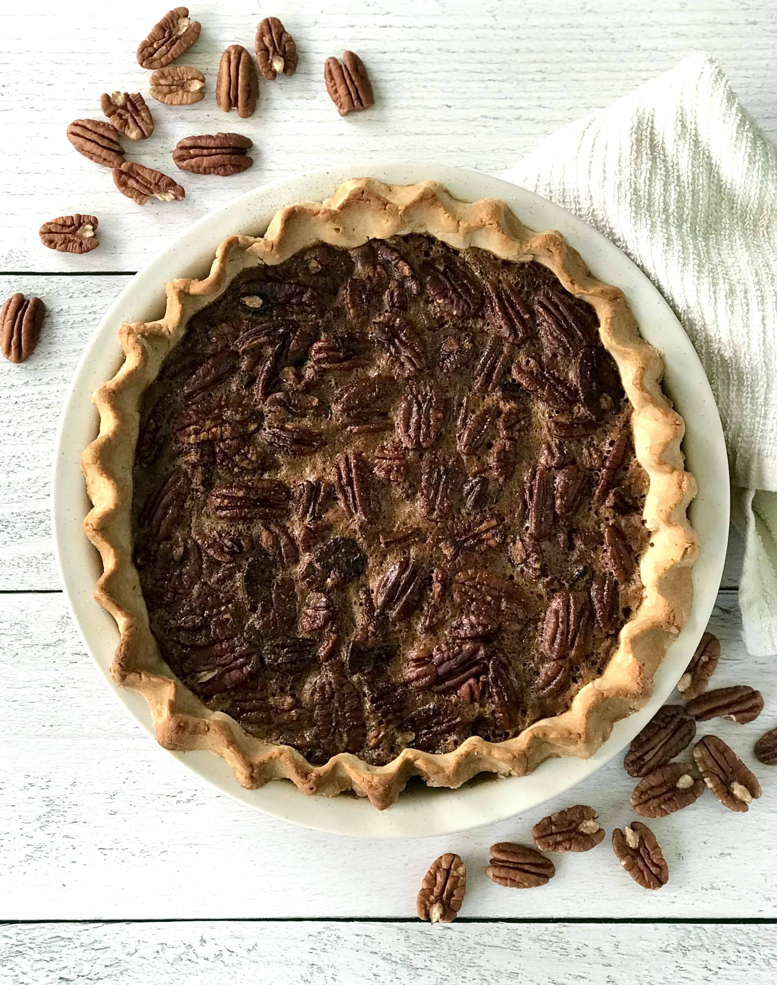 Dairy-free pecan pie filling in a grain-free crust in a pie dish on a white wooden table strewn with pecans.