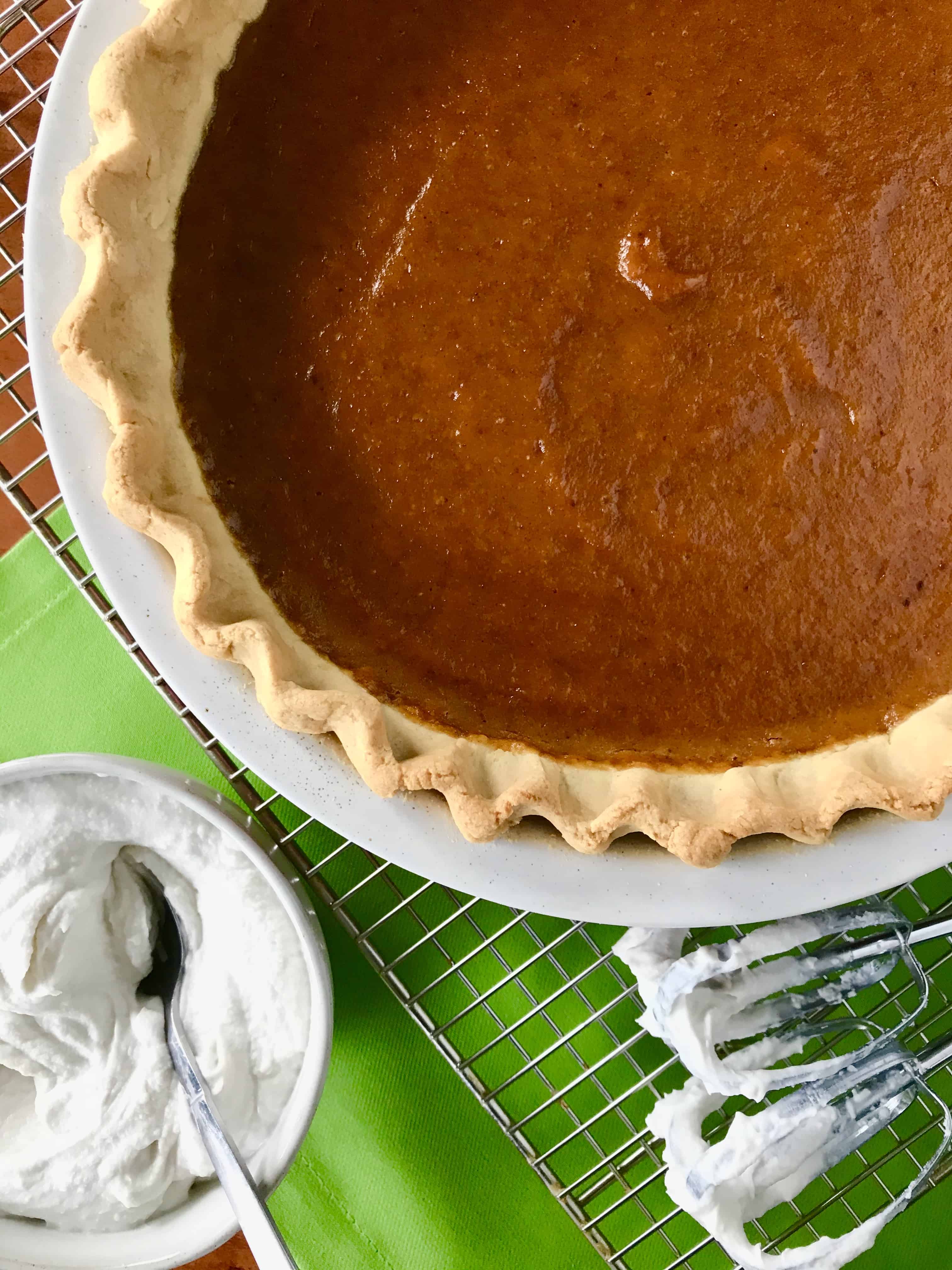 A Paleo pumpkin dessert in a pie dish on a cooling rack next to a bowl of whipped coconut cream.