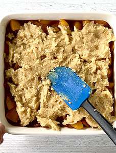 A spatula smoothing spoonfuls of cobbler topping batter over chopped peaches in a white square baking dish.