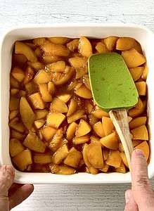 A hand holding a white square baking dish while the other hand holds a spatula smoothing chopped peaches into an even layer in the dish.