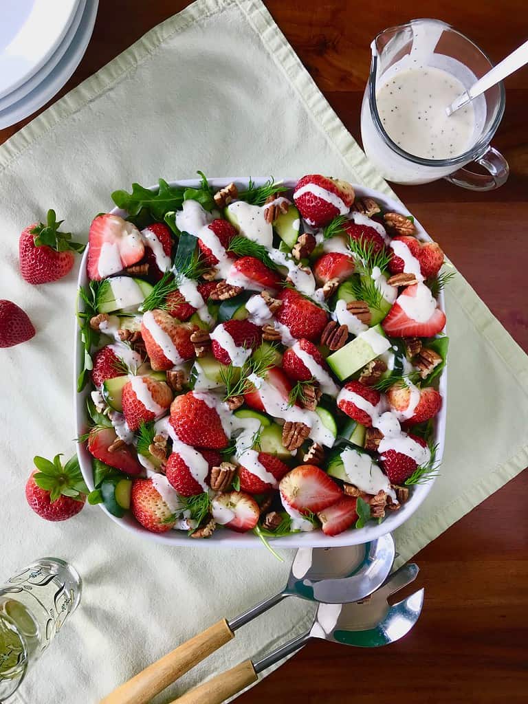 Arugula salad with strawberries, pecans and a yogurt dressing in a white square bowl next to salad tongs and glass pitcher of more dressing.