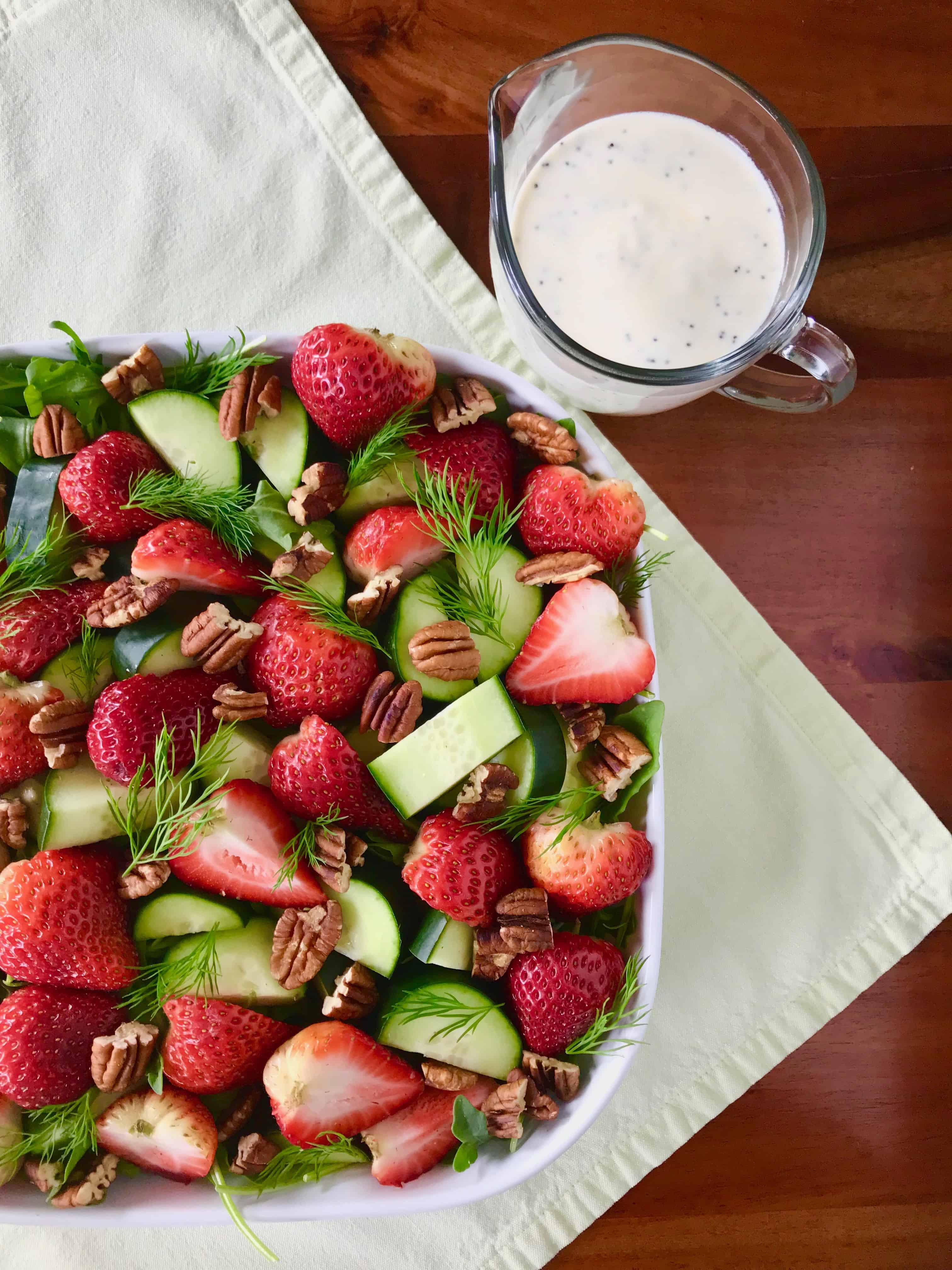 Healthy arugula salad with berries and nuts in a white square bowl next to a glass pitcher filled with a creamy dressing.