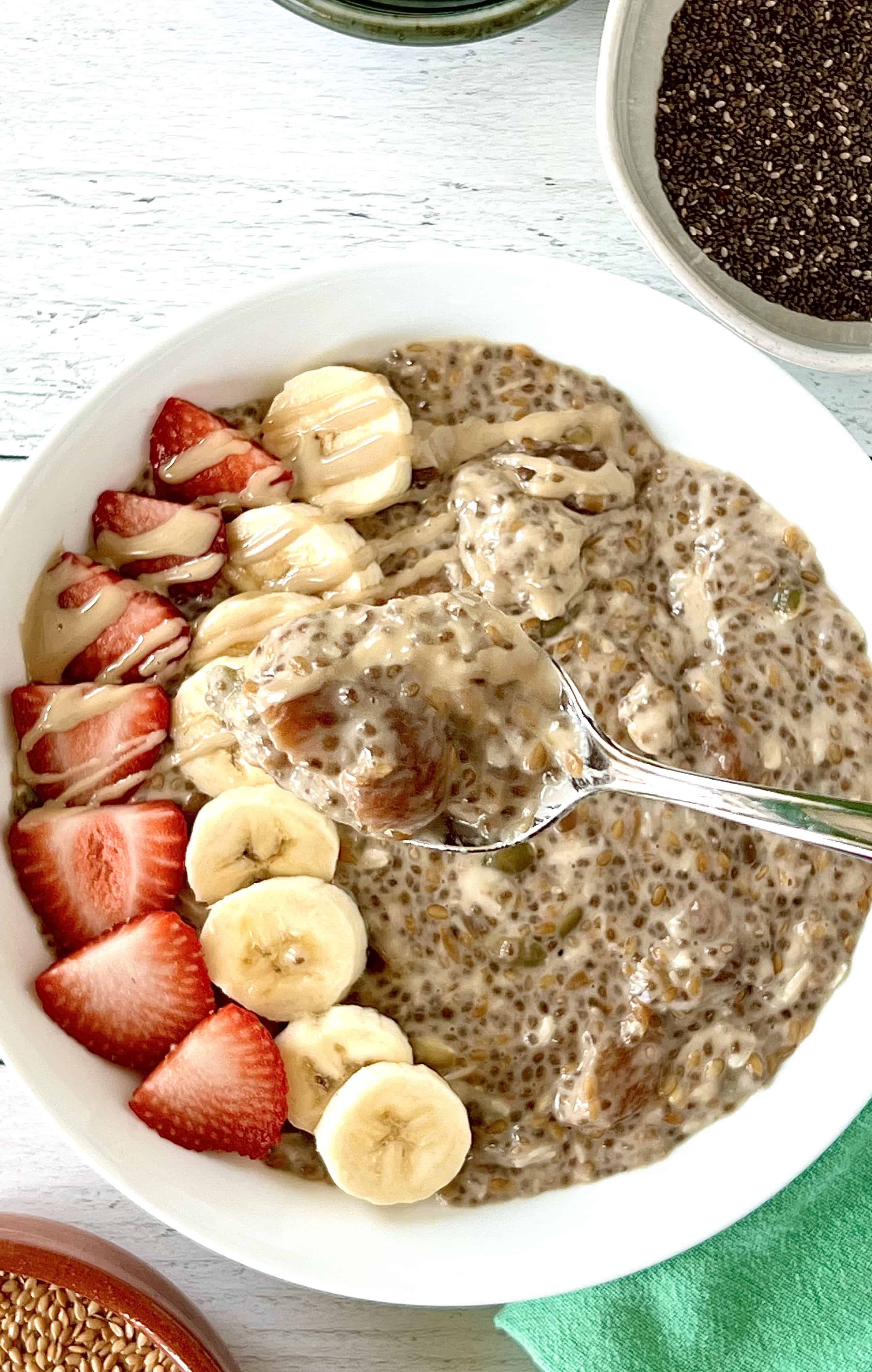 Grain-free Paleo oatmeal made with seeds and non-dairy milk in a shallow white bowl topped with sliced strawberries and bananas with spoon holding some pudding up over the bowl.