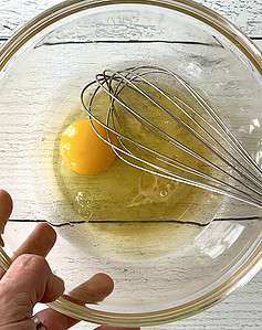 A hand holding a glass bowl containing the wet ingredients for a cobbler topping and a whisk.