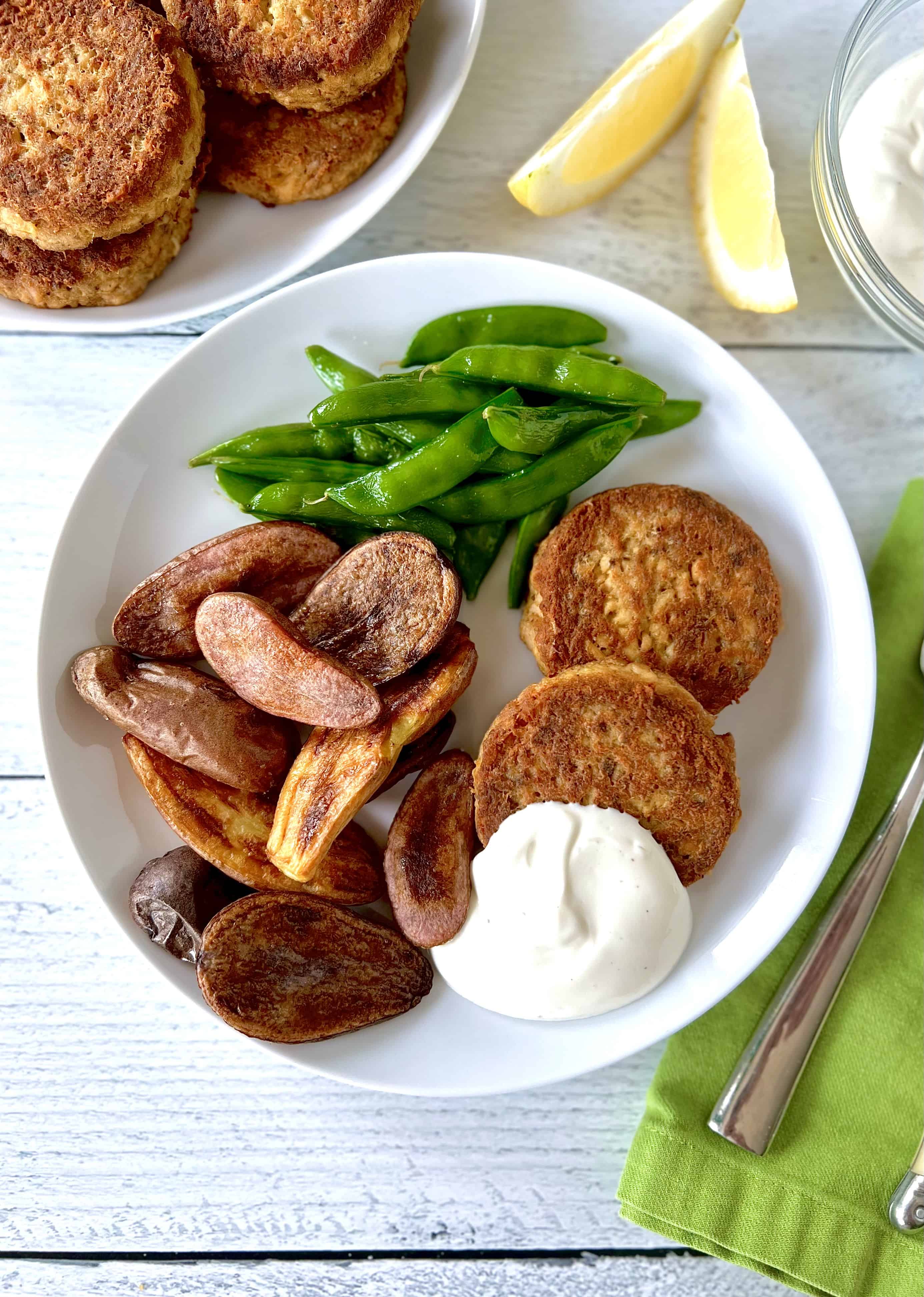 Healthy salmon patties on a white plate with a dollop of lemon aioli, roasted potatoes and steamed sugar snap peas.