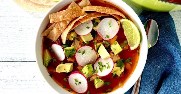 Whole30 chicken tortilla soup in a white bowl topped with sliced radishes, chopped parsley, cubed avocado, crispy tortilla strips and a lime wedge.