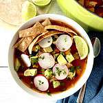 Healthy tortilla soup in a white bowl topped with sliced radishes, cubed avocado, chopped parsley, crispy tortilla strips and a lime wedge.