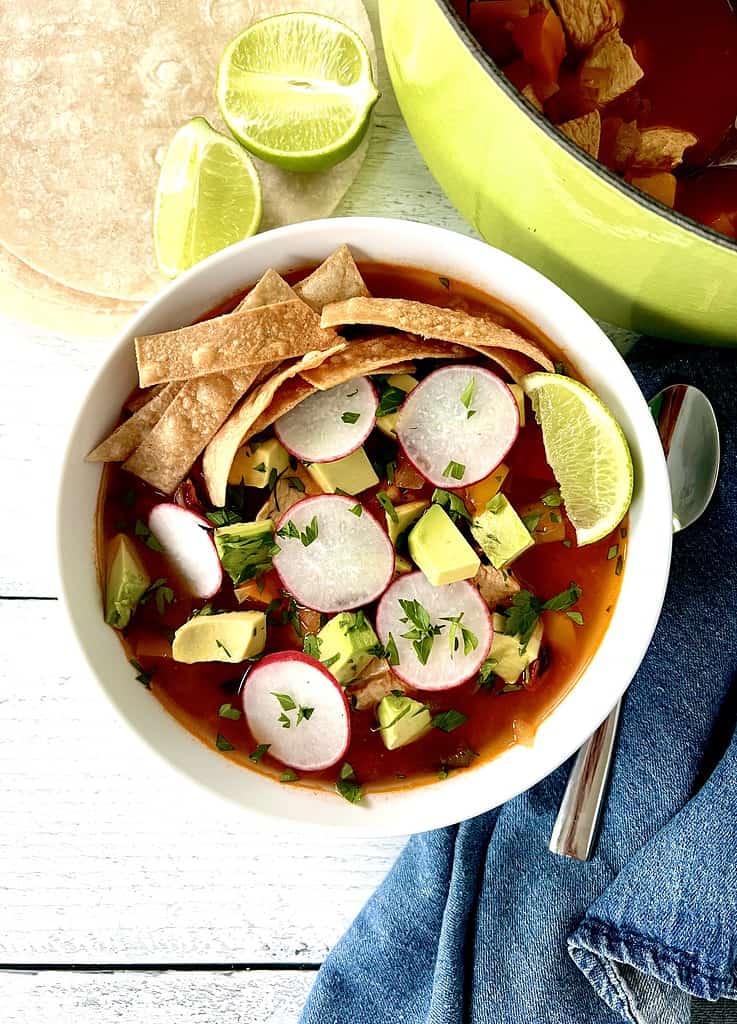 Healthy tortilla soup in a white bowl topped with sliced radishes, cubed avocado, chopped parsley, crispy tortilla strips and a lime wedge.