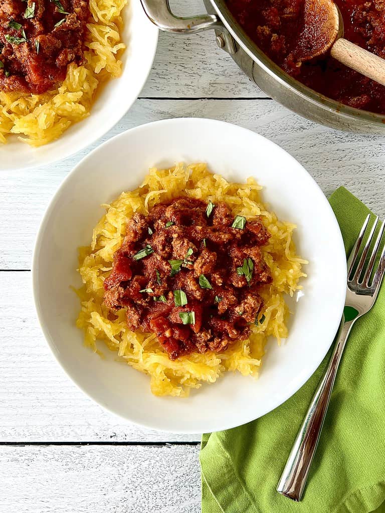 Spaghetti squash topped with ground beef in a tomato sauce in a white bowl next to a green napkin with a fork on it.