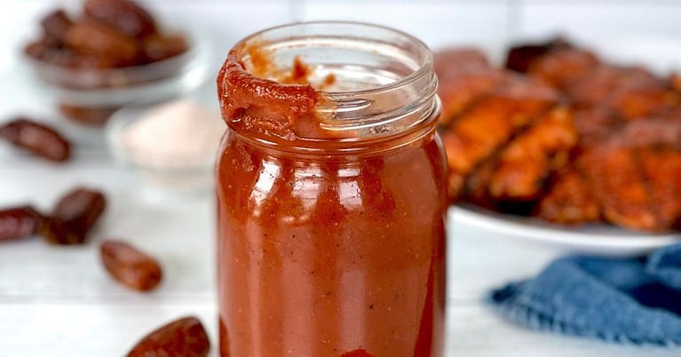 Healthy homemade barbecue sauce in a glass jar in front of a plate of BBQ grilled chicken.
