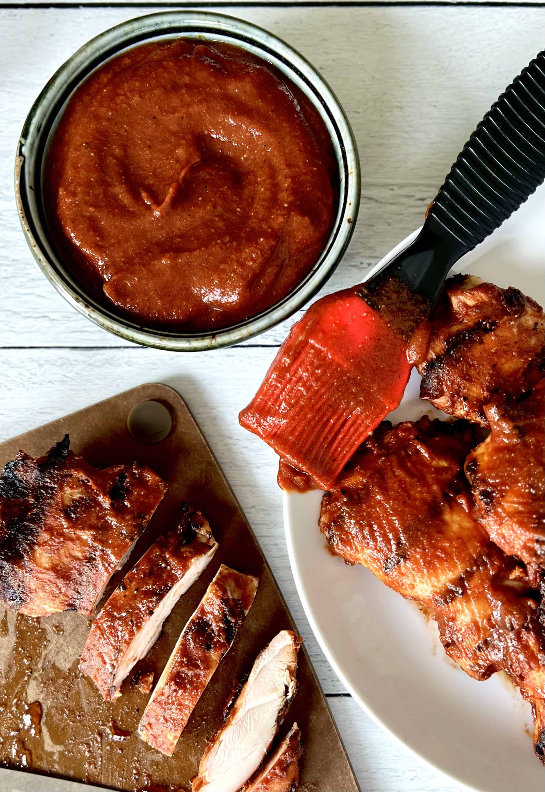 Healthy BBQ sauce in a bowl next to a basting brush coated in the sauce resting on the rim of a white plate with BBQ chicken on it and a cutting board with sliced BBQ chicken on it.