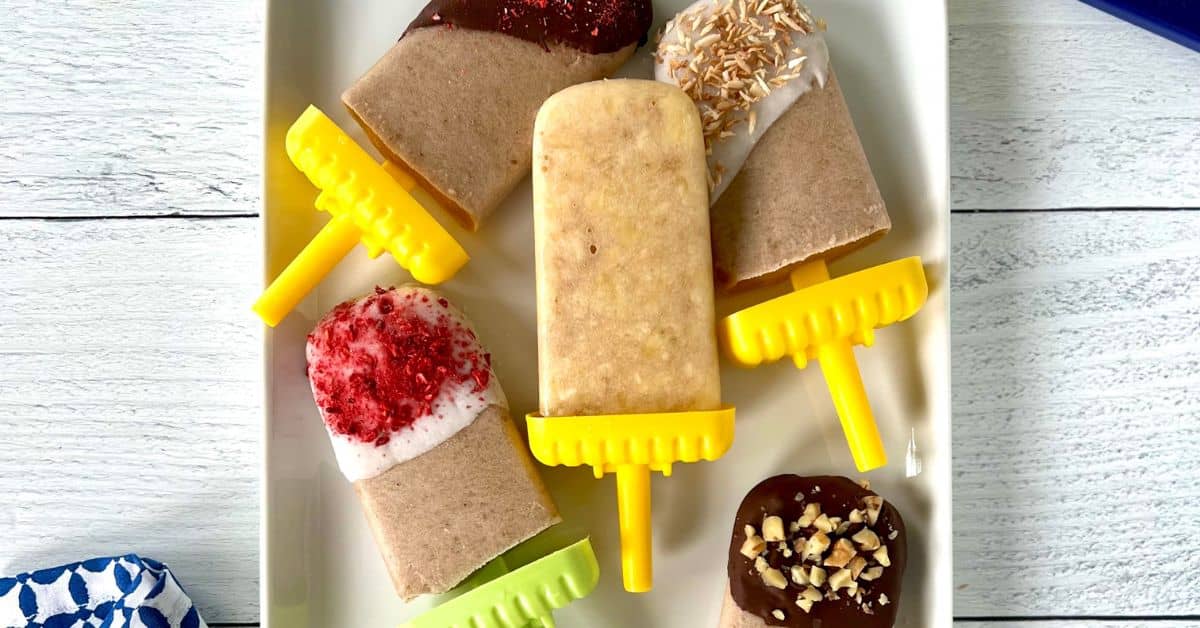 Banana ice cream pops on a white rectangular platter - some covered in chocolate, some in yogurt, all with different toppings.