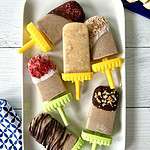 Frozen banana pops on a white rectangular platter, some dipped in chocolate, some in yogurt, all with different toppings.
