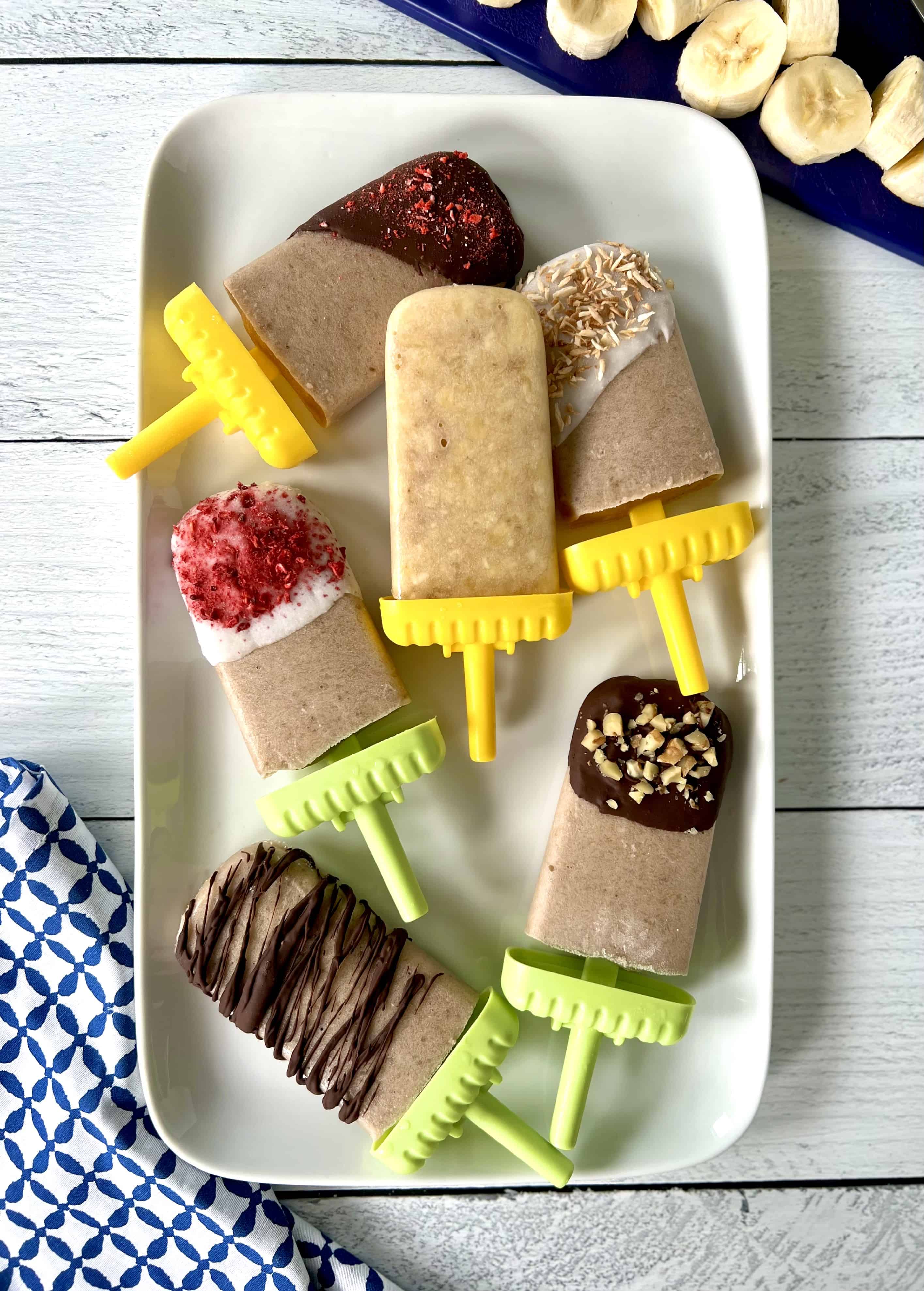 Frozen banana pops on a white rectangular platter, some dipped in chocolate, some in yogurt, all with different toppings.