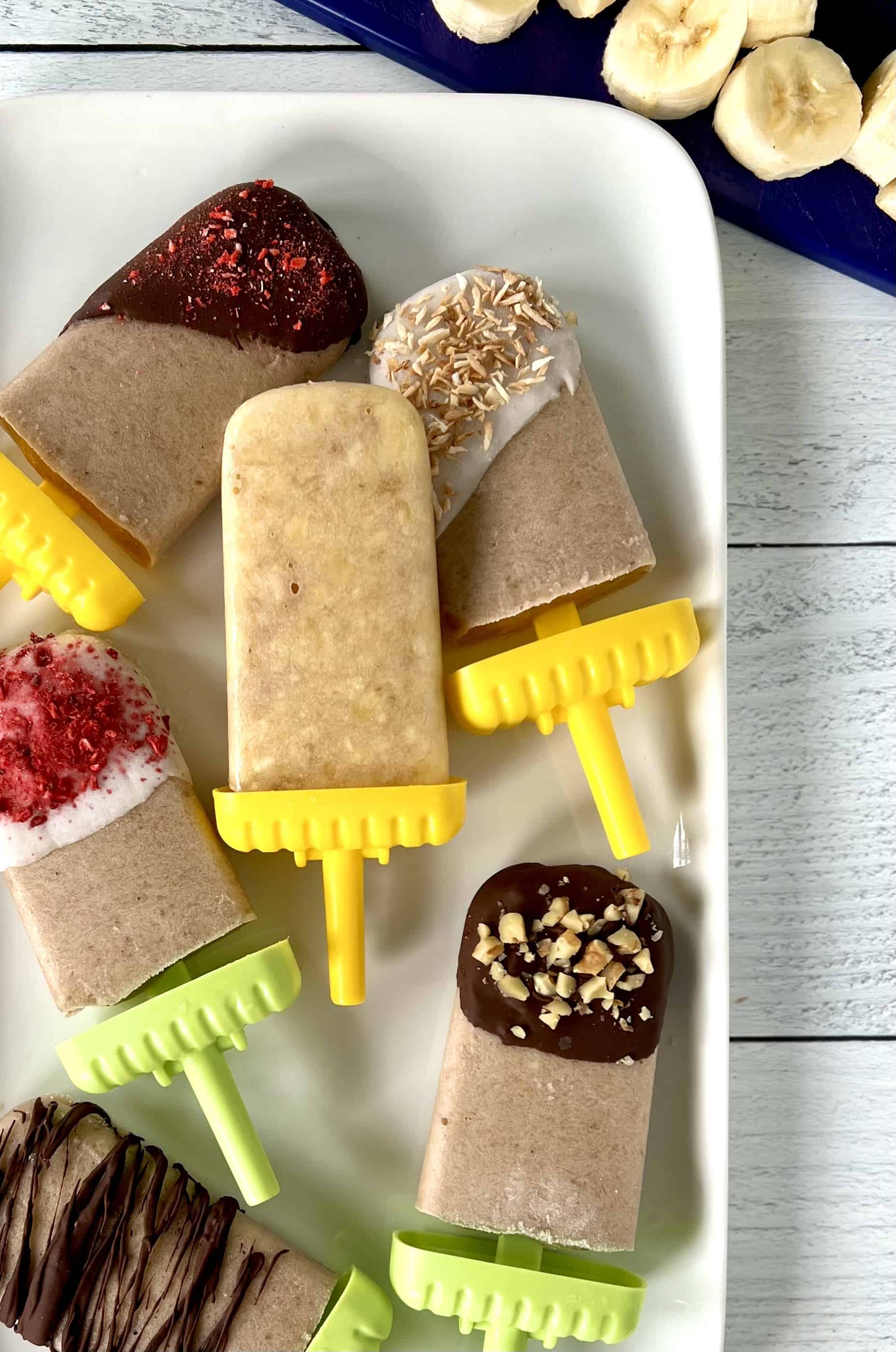 Healthy homemade popsicles on a white rectangular platter, some dipped in chocolate, some in yogurt, all with different toppings.