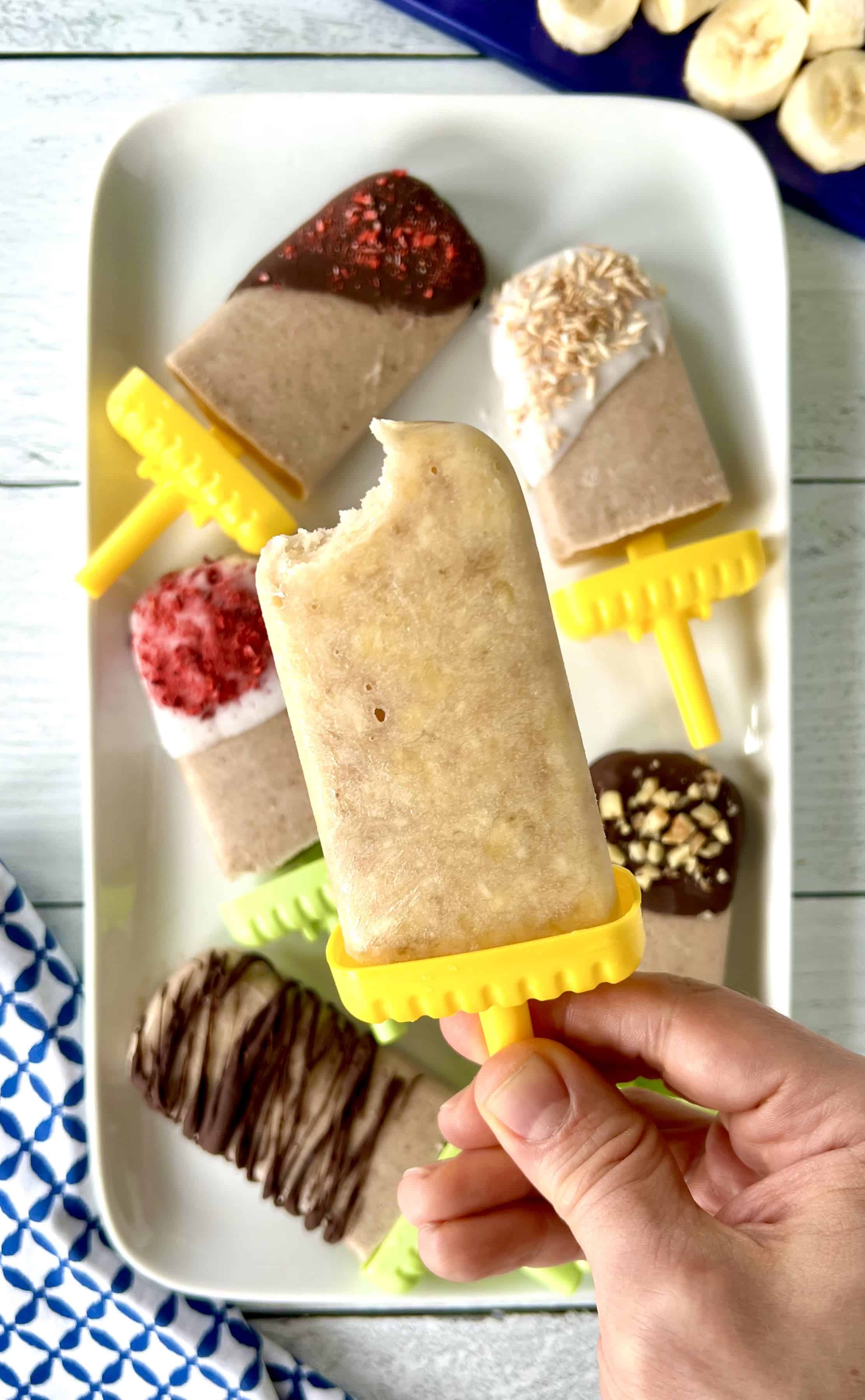 Homemade frozen popsicles on a white platter, some dipped in chocolate, some in yogurt, all with different toppings, and a hand holding a plain one above the others.