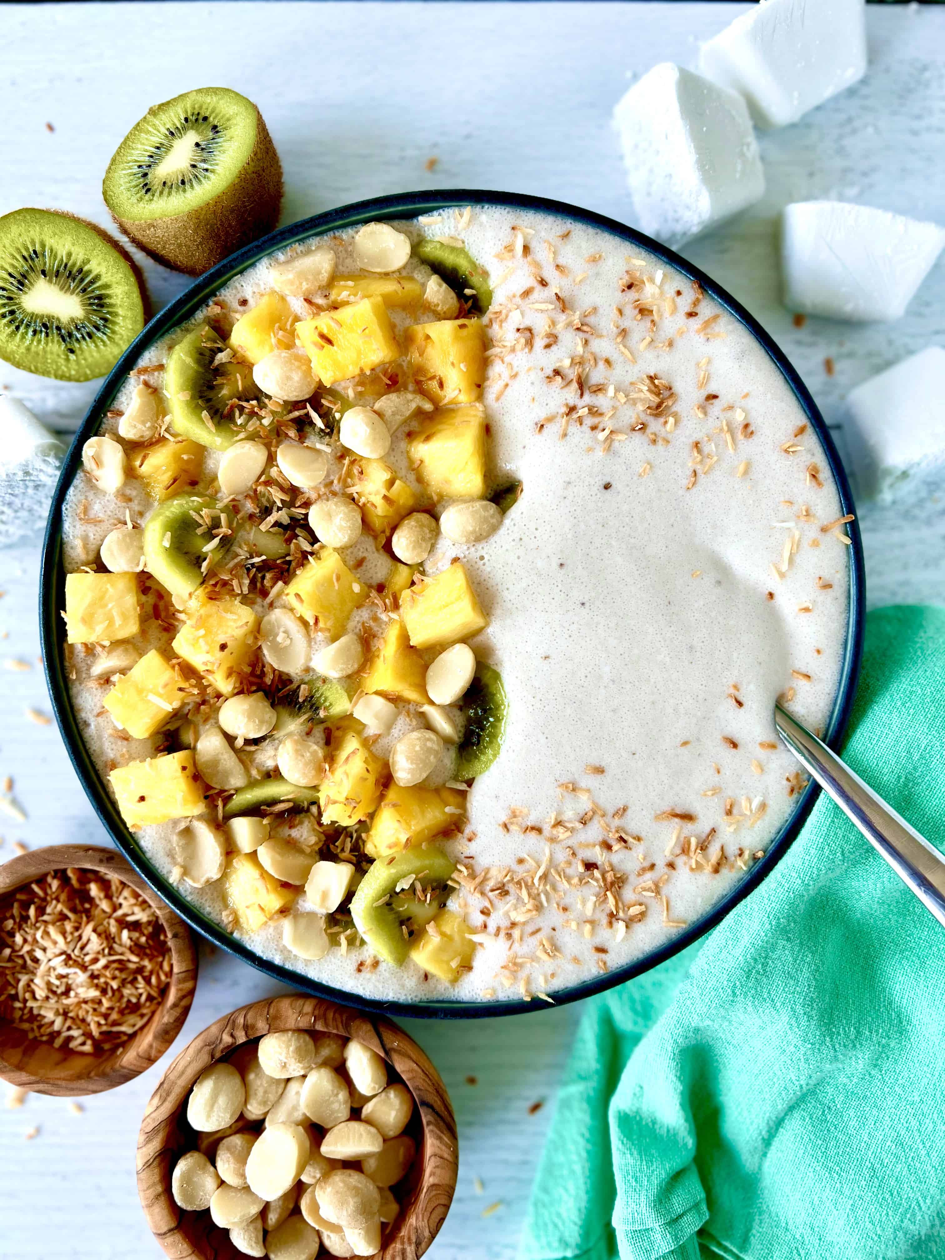 Paleo smoothie bowl made with frozen bananas, topped with pineapple, kiwi, macadamias and toasted shredded coconut, next to bowls of more macadamias and shredded coconut and a halved kiwi.