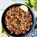 A Paleo cauliflower rice dish in a blue bowl surrounded by parsley, limes, a blue patterned napkin and a spoon.