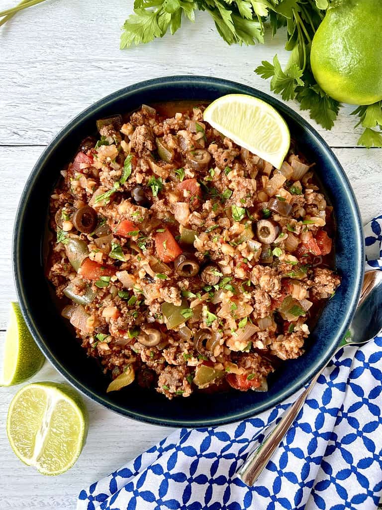 A Paleo cauliflower rice dish in a blue bowl surrounded by parsley, limes, a blue patterned napkin and a spoon.