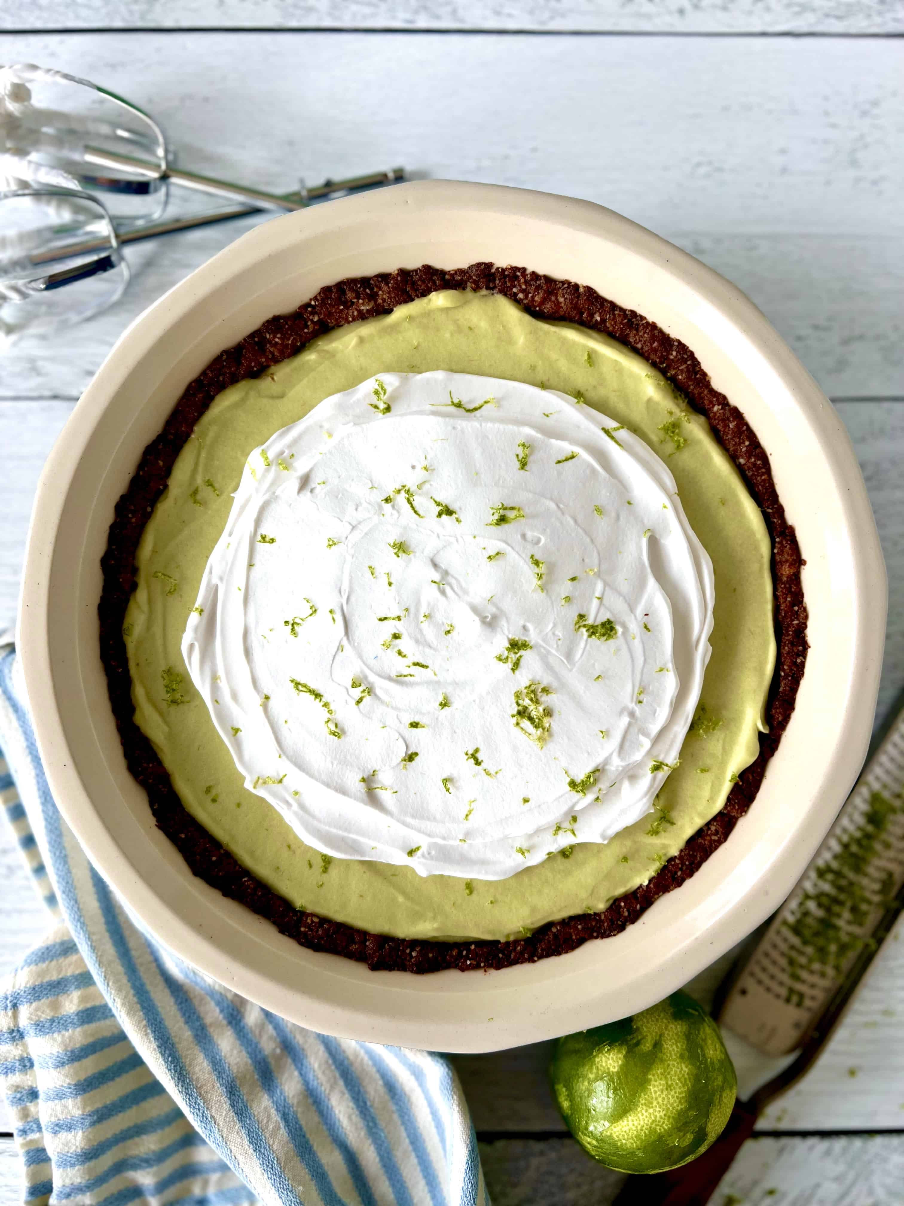 Gluten-free key lime pie in a pie dish next to a blue and white-striped towel, a lime, a lime zester and hand mixer beaters covered in whipped coconut cream.