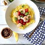 Whole30 Shrimp Scampi with spaghetti squash and cherry tomatoes in a white bowl next to a blue and white patterned napkin with a fork on it.