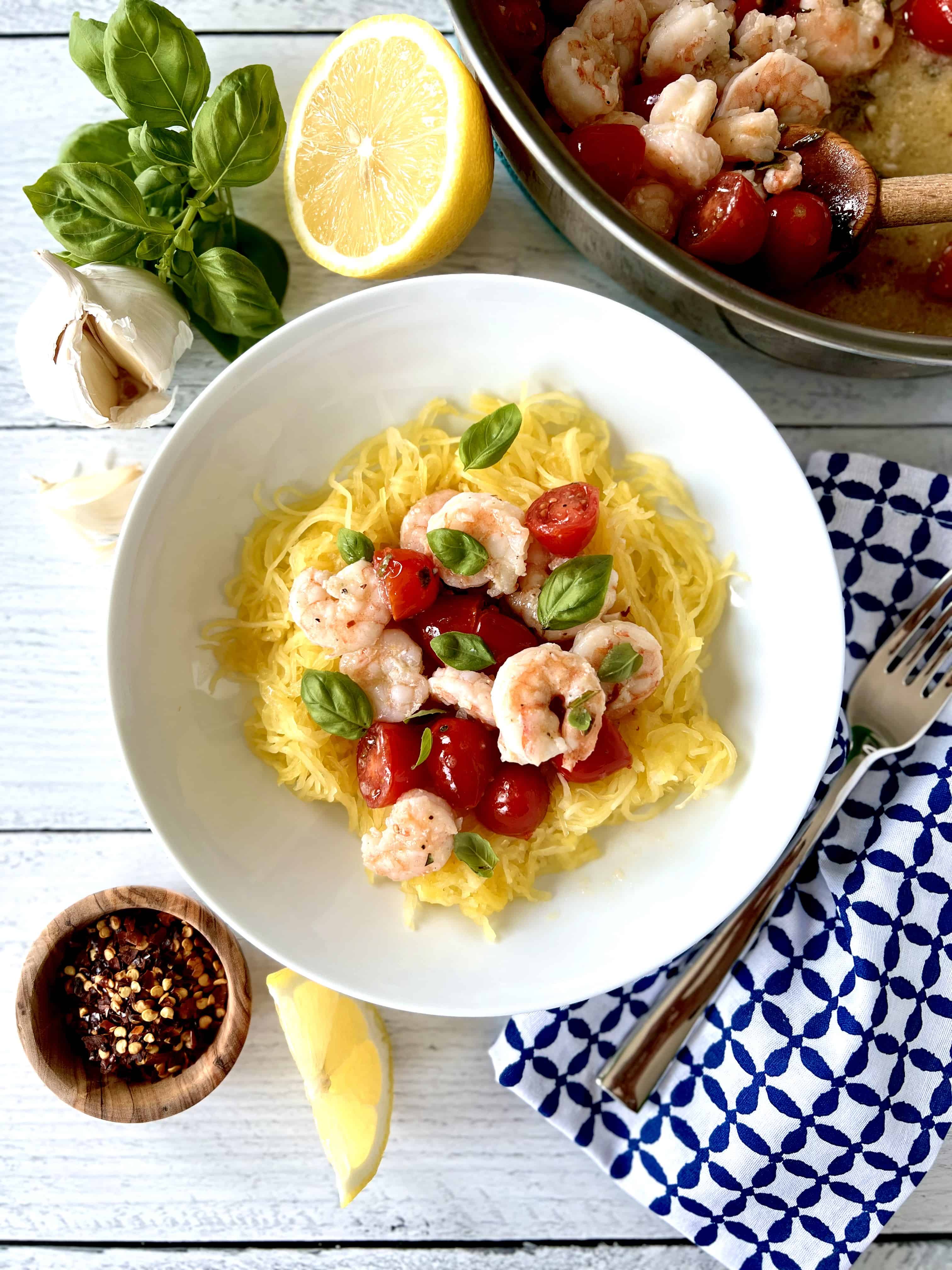Whole30 Shrimp Scampi with spaghetti squash and cherry tomatoes in a white bowl next to a blue and white patterned napkin with a fork on it.