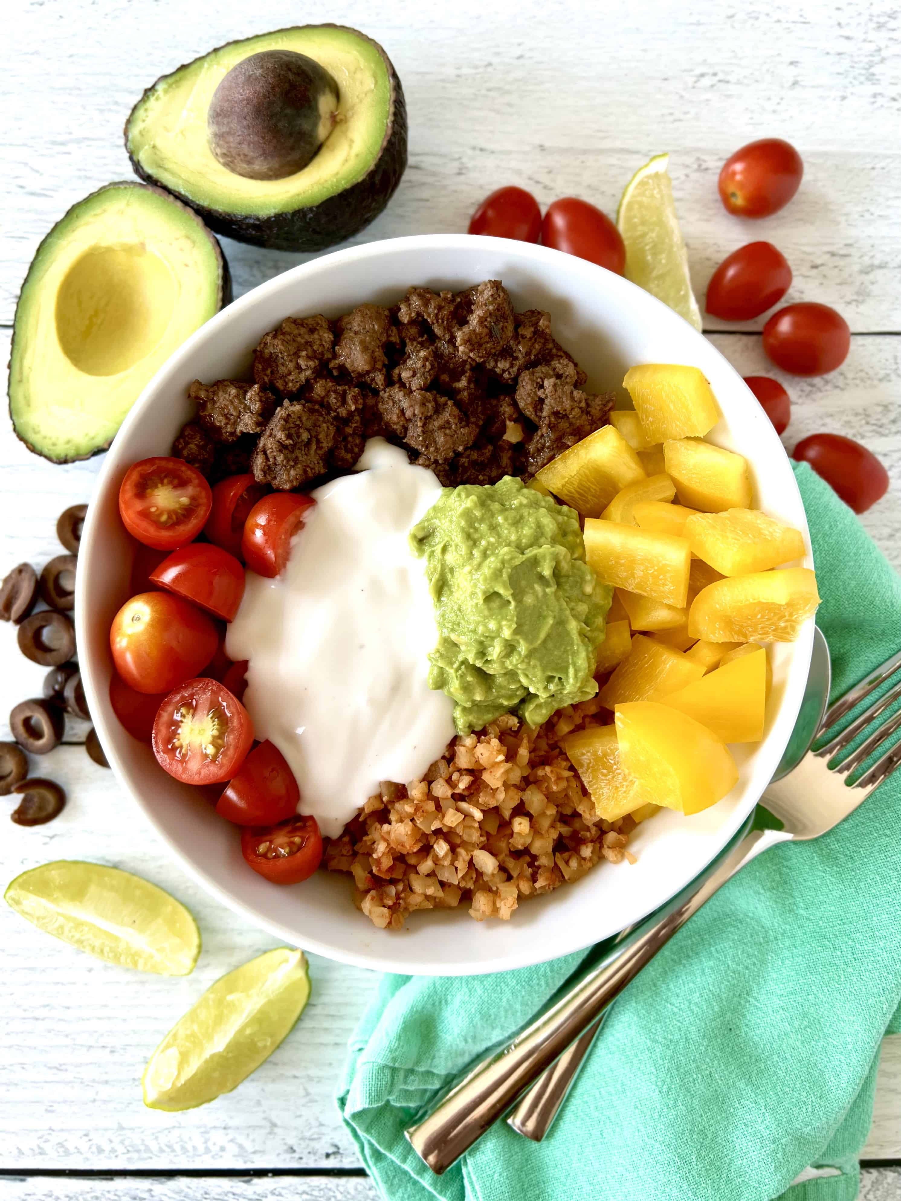 A low carb taco bowl on a white wooden table surrounded by lime wedges, avocado halves, cherry tomatoes and sliced black olives.