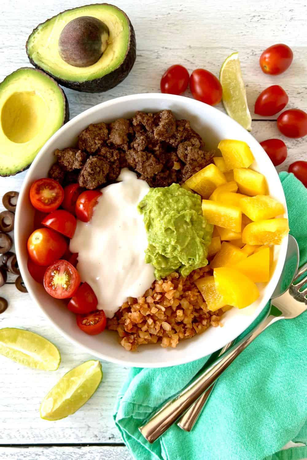 A Whole30 taco bowl on a white wooden table surrounded by avocados, cherry tomatoes, lime wedges and sliced black olives.