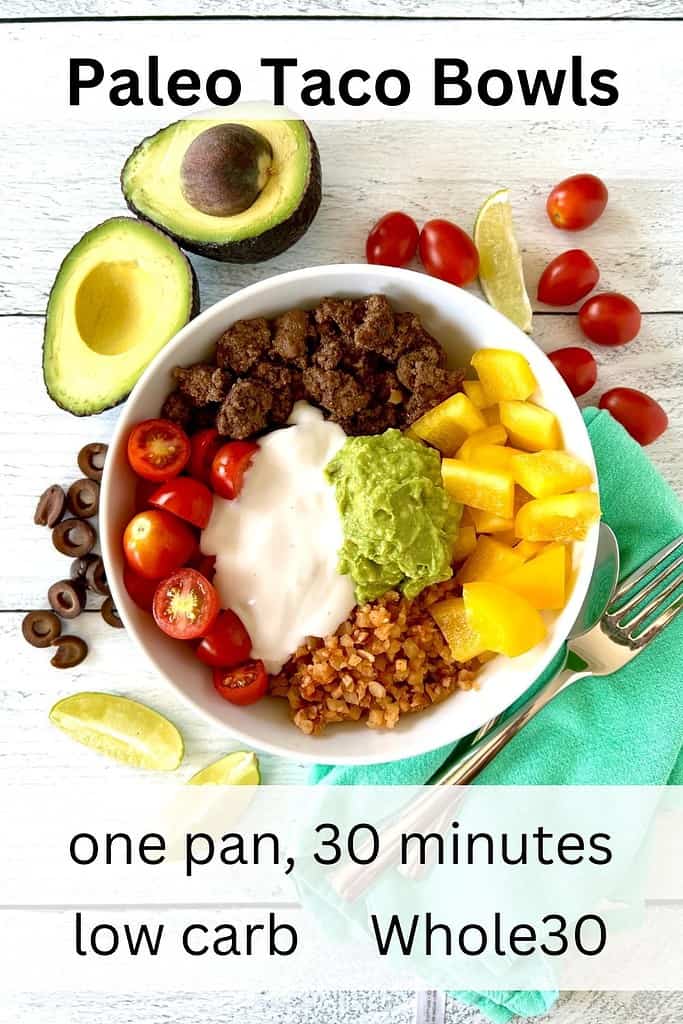 A Paleo taco bowl on a white wooden table surrounded by lime wedges, sliced black olives, cherry tomatoes and halved avocados.