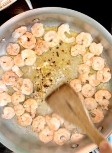 Stirring garlic and red pepper flakes into shrimp in a large stainless steel pan.