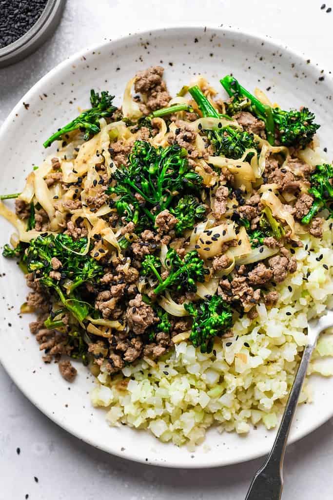 Ground beef, cabbage and broccolini on cauliflower rice on a white speckled plate.