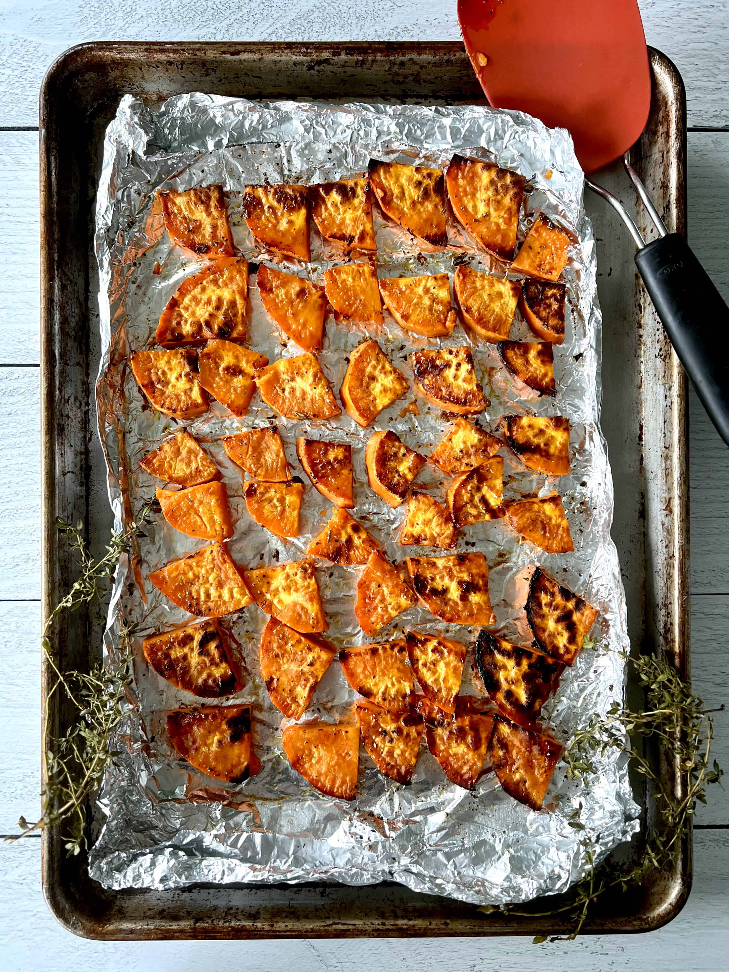 Slightly charred slices of sweet potato on a foil-lined baking sheet with a spatula and thyme sprigs.