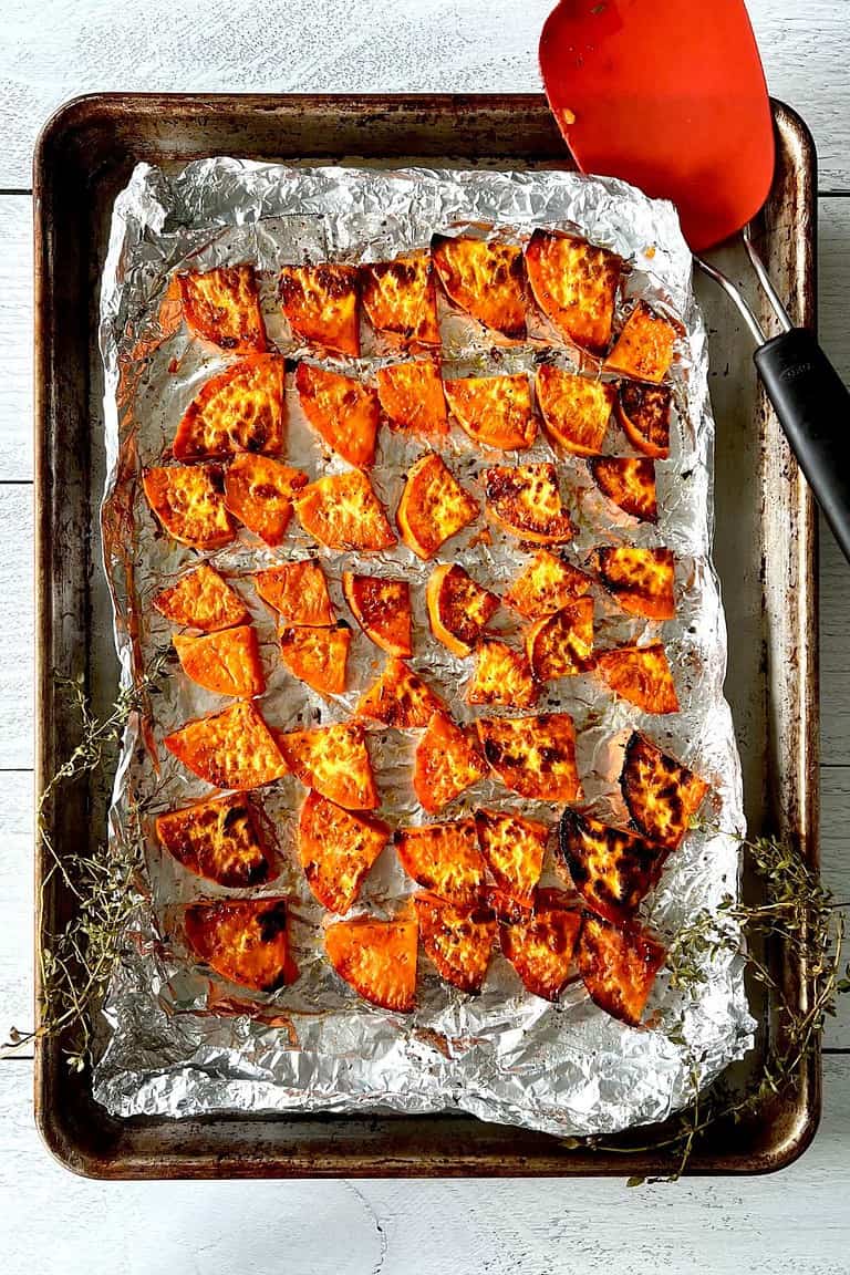 Slices of caramelized sweet potatoes on a foil-lined baking sheet with a spatula and thyme sprigs.