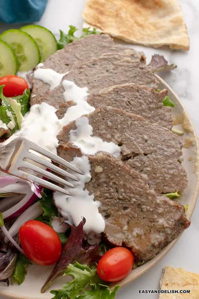 Sliced gyro meat drizzled with a creamy sauce next to cucumbers and cherry tomatoes on a plate.