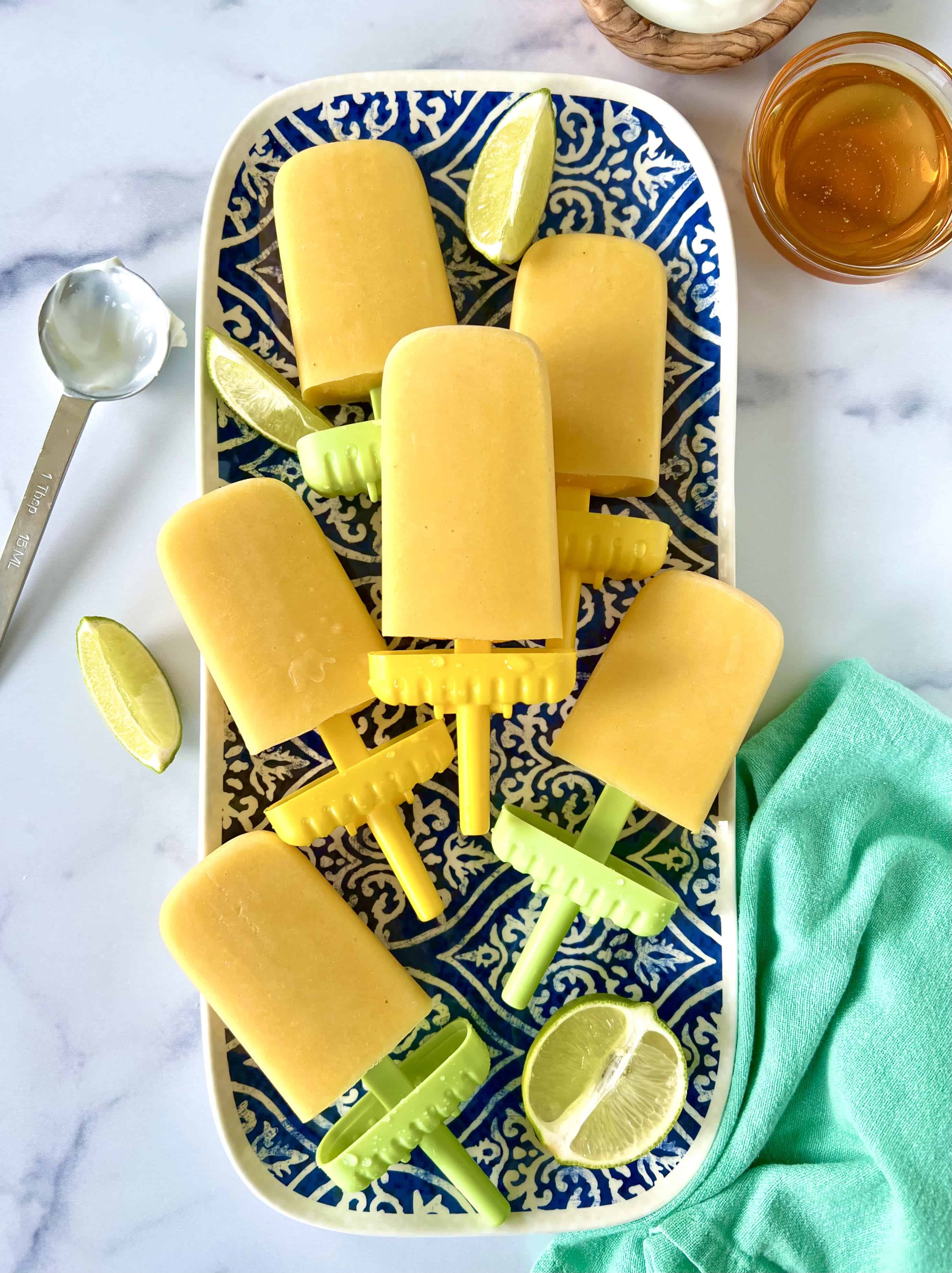 Mango, yogurt, honey and lime popsicles on a blue and white patterned platter with lime wedges.