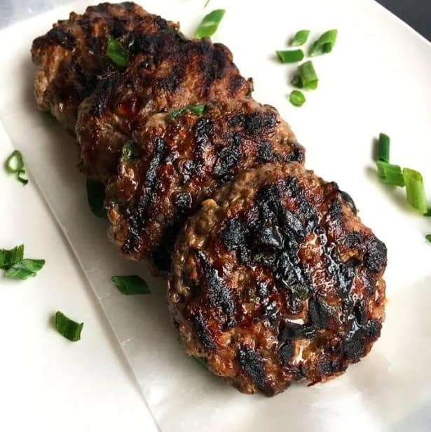 4 beef burger patties lined up on a white platter strewn with sliced green onions.