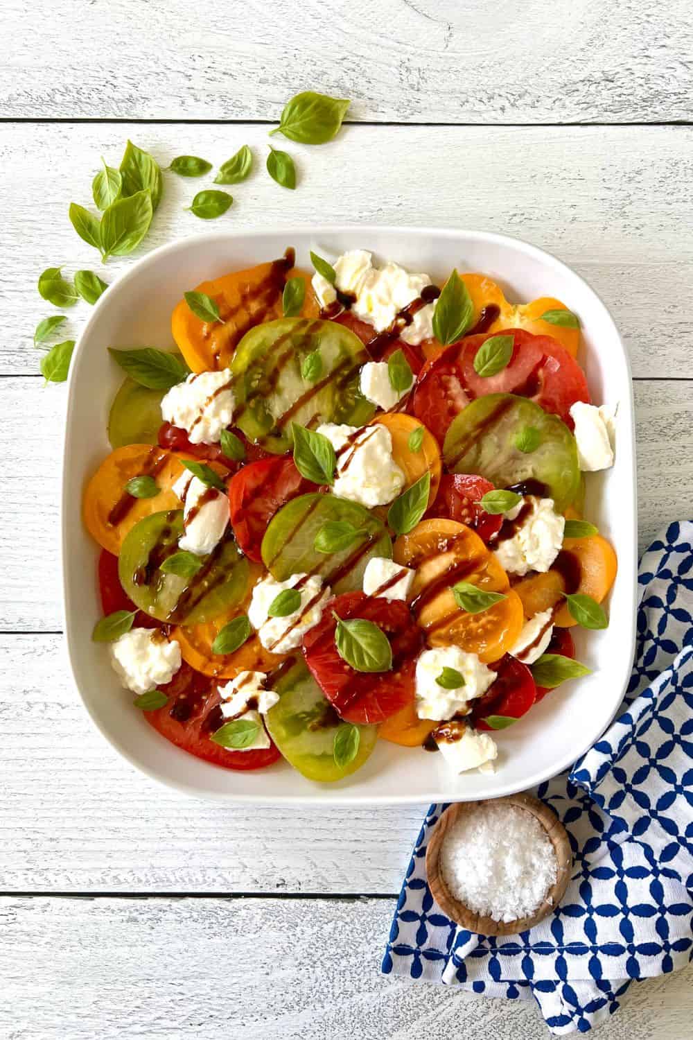 A tomato salad with burrata cheese and basil leaves in a white platter.