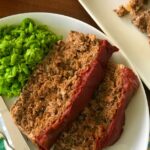 A no bread crumbs meatloaf sliced on a white plate with mashed peas.