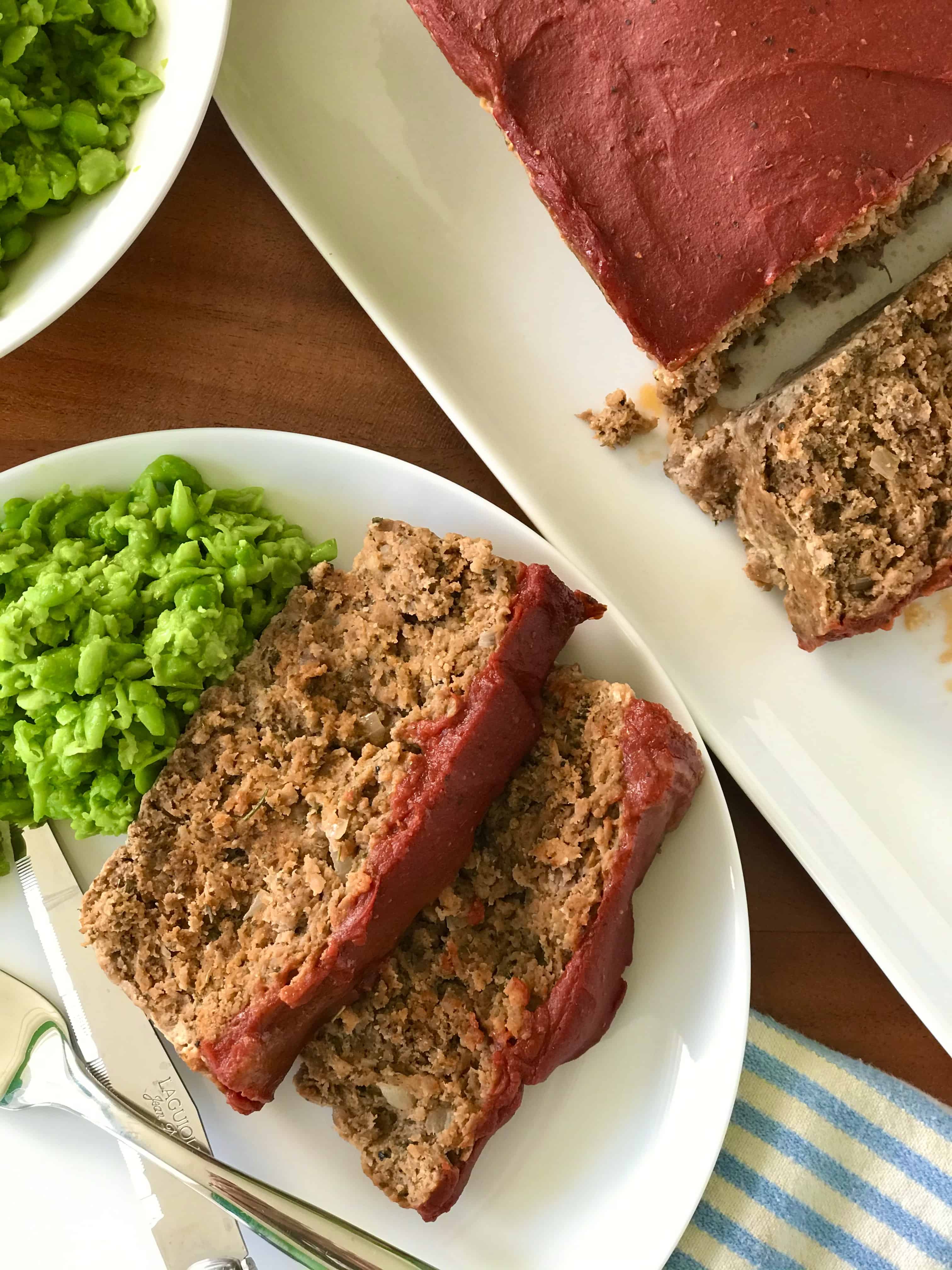 A Paleo meatloaf sliced on a white plate with mashed peas, fork and knife.