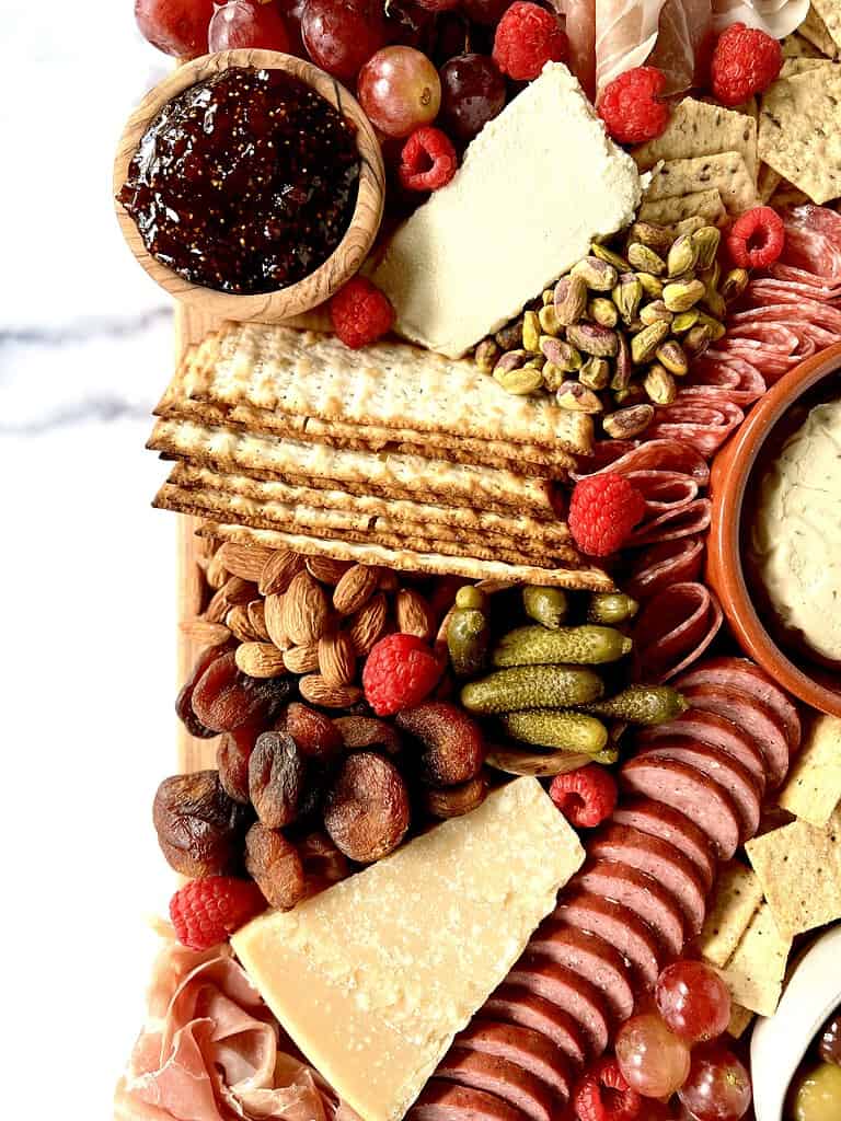 A Gluten-Free Cheese Board with meats, crackers, olives, almonds, pistachios, raspberries, grapes, dried apricots and cornichons on a wooden cutting board on a marble table.