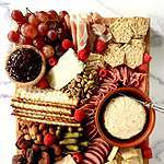 A gluten-free party platter cheeses, meats, crackers, olives, nuts, fruit, dried fruit and cornichon on a wooden cutting board on a marble table.