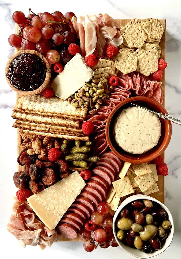 A gluten-free party platter cheeses, meats, crackers, olives, nuts, fruit, dried fruit and cornichon on a wooden cutting board on a marble table.