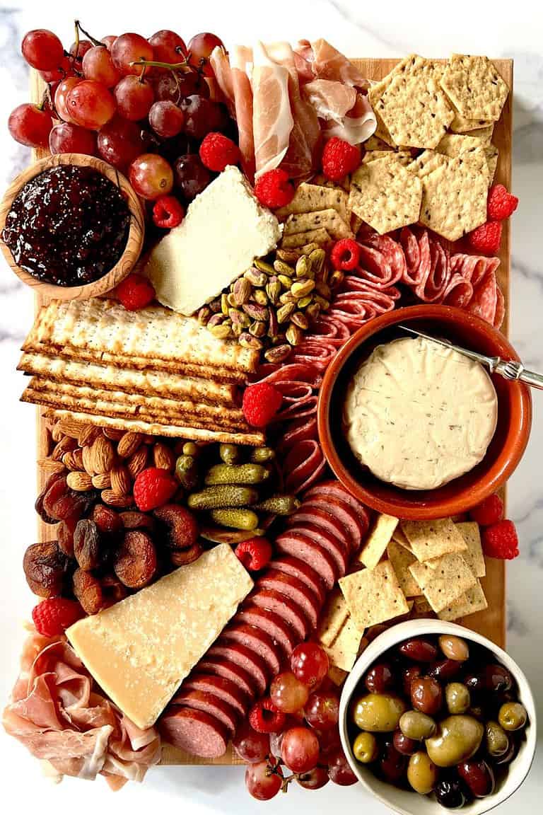A Charcuterie Board with cheeses, meats, glute-free crackers, olives, nuts, fruit, dried fruit and cornichon on a wooden cutting board on a marble table.