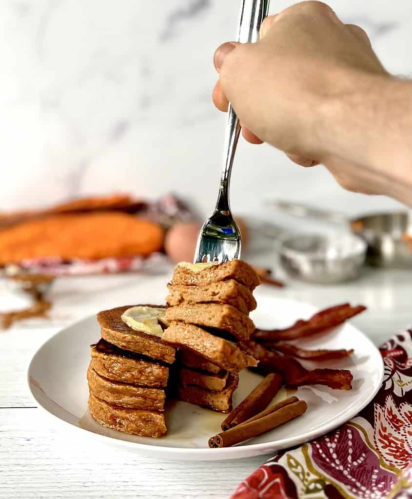 Grain-free pancakes stacked on a plate with a hand using a fork to lift out a stack of bites.