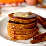 Paleo Sweet Potato Pancakes stacked on a white plate topped with ghee and maple syrup, next to cinnamon sticks and bacon.