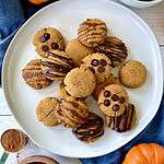 Vegan pumpkin cookies piled on a plate, some with chocolate chips, some with a chocolate or caramel drizzle, some plain.